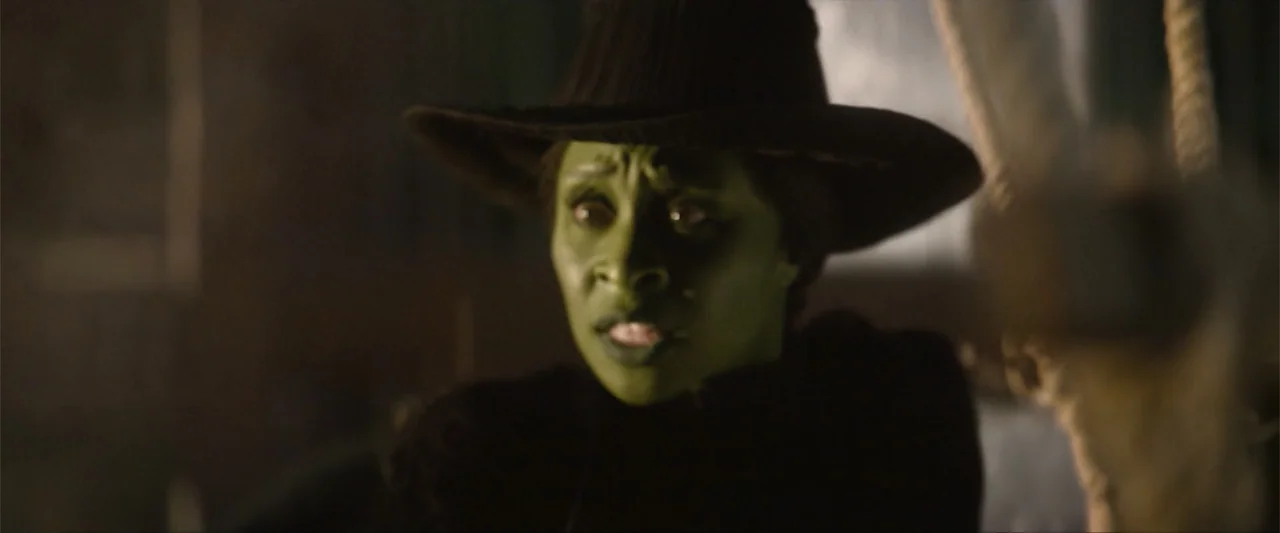 Wicked (1998) movie still: closeup — A dimly lit interior space contains a person with green skin and a hat. The individual gazes forward with a…