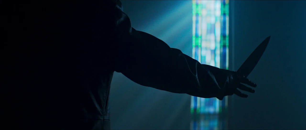 The Sacrifice Game (2023) movie still: closeup — A darkened interior space features a tall, narrow window with blue stained glass panels. A…