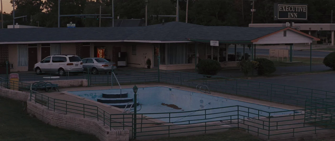 Mud (2012) movie still: establishing — An exterior view of a low-rise motel with an empty, drained swimming pool. A dark parking lot features two…