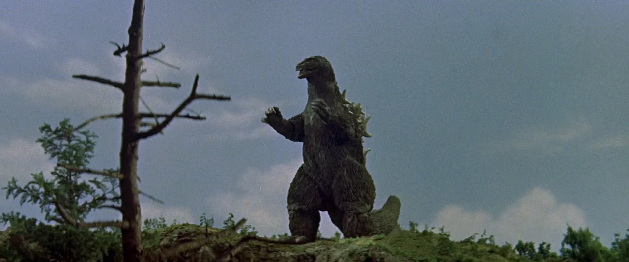 King Kong Vs. Godzilla (1962) movie still: fullbody — A miniature outdoor set features a reptilian creature standing atop a grassy mound. A leafless…