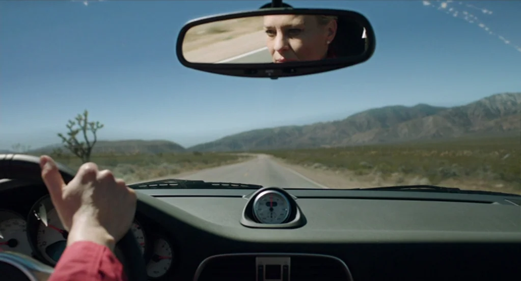 The Congress (2013) movie still: wide — The view from a car dashboard shows a desolate road stretching through a desert. A blonde woman driving is…