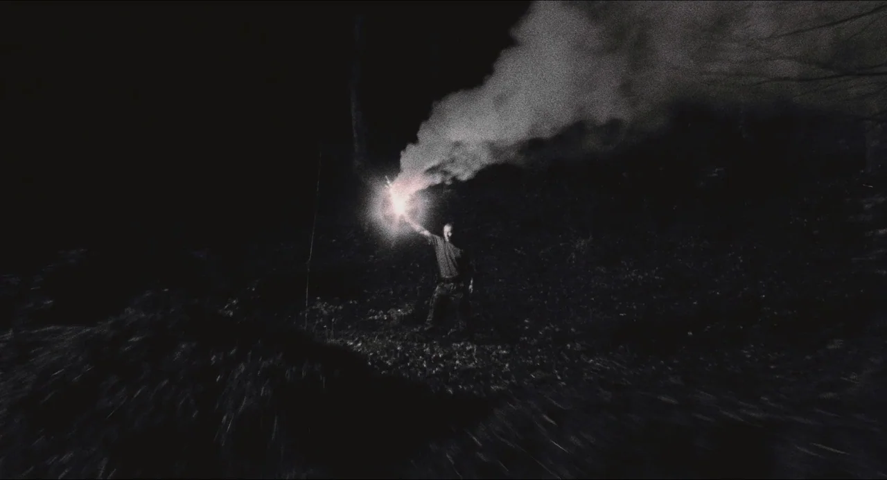 Dog Soldiers (2002) movie still: wide — A dark wooded area is illuminated by a single handheld signal flare. A man stands at center frame holding…
