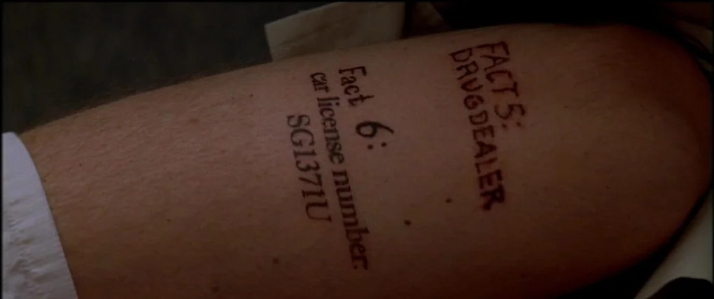 Memento (2000) movie still: closeup — A close-up view shows a portion of a person's upper arm. Tattoos are visible on the skin consisting of text…