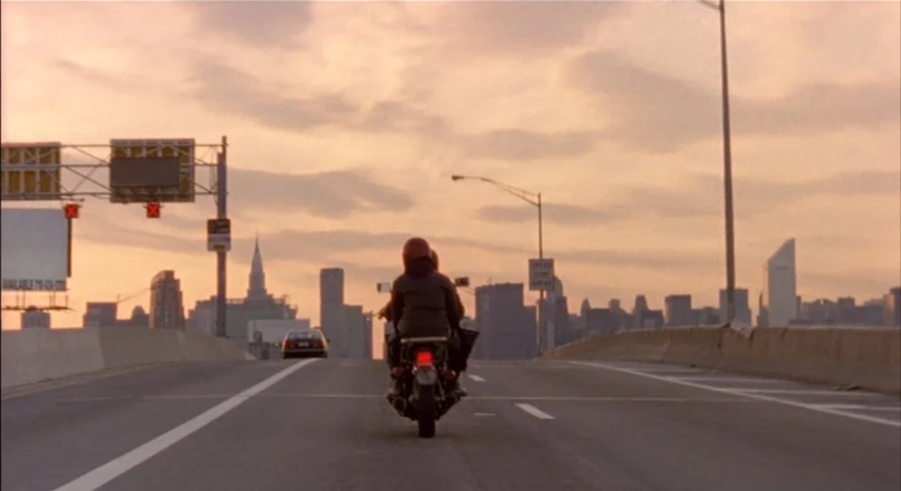 Hamlet (2000) movie still: wide — A wide highway stretches toward a distant urban skyline under a warm, cloudy sunset. Two people on a motorcycle…