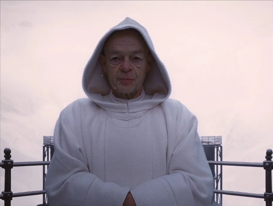 The Grand Budapest Hotel (2014) movie still: medium — A man in a white robe stands on a snowy mountain viewing platform. Metal railings flank the…