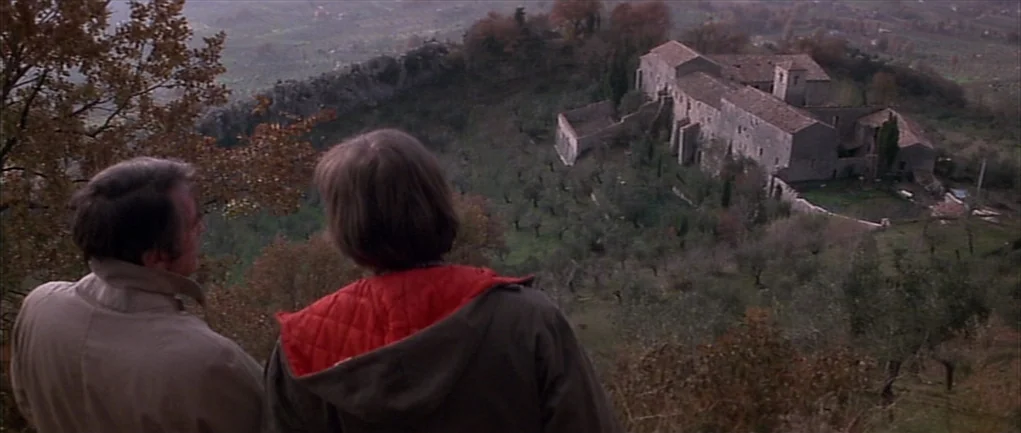 The Omen (1976) movie still: wide — An Italian hillside landscape features a remote stone building nested among olive trees. Two men stand on a…