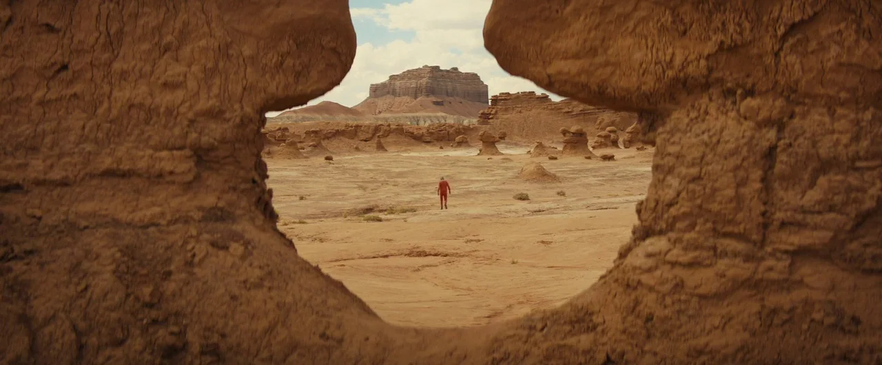 Damsel (2018) movie still: wide — A wide desert landscape featuring a large, weathered rock formation framing the center view. A solitary figure…