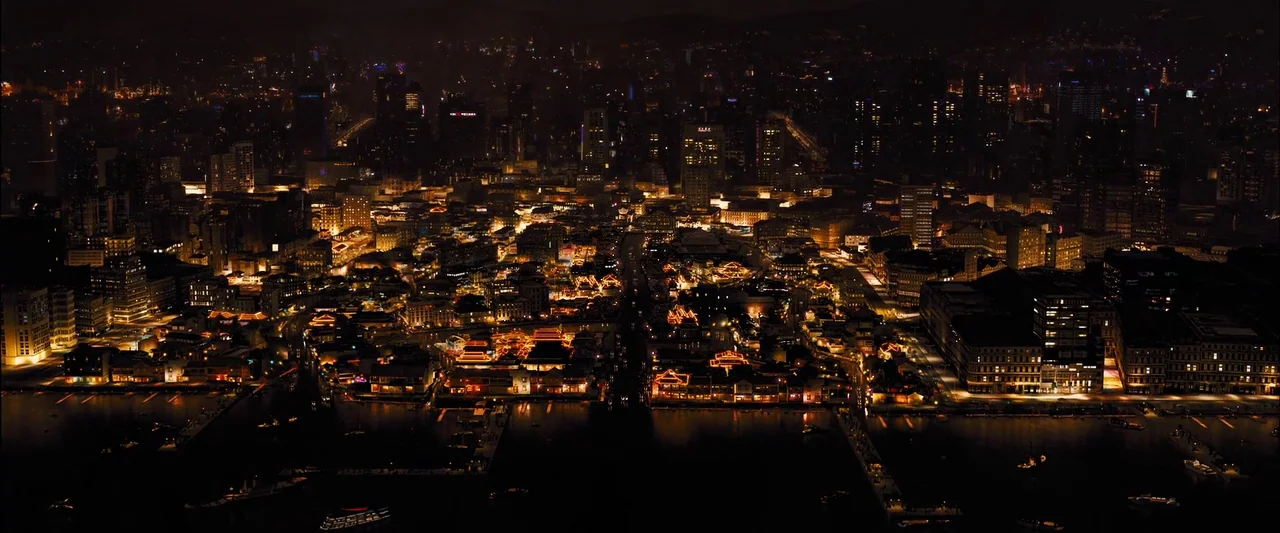 Skyfall (2012) movie still: establishing — A high-angle view captures a dense urban city at night bordering a dark river. Numerous illuminated…