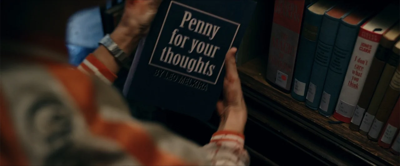 Gunpowder Milkshake (2021) movie still: closeup — A person holds a dark blue book with white text in a library setting. Several colorful book spines…
