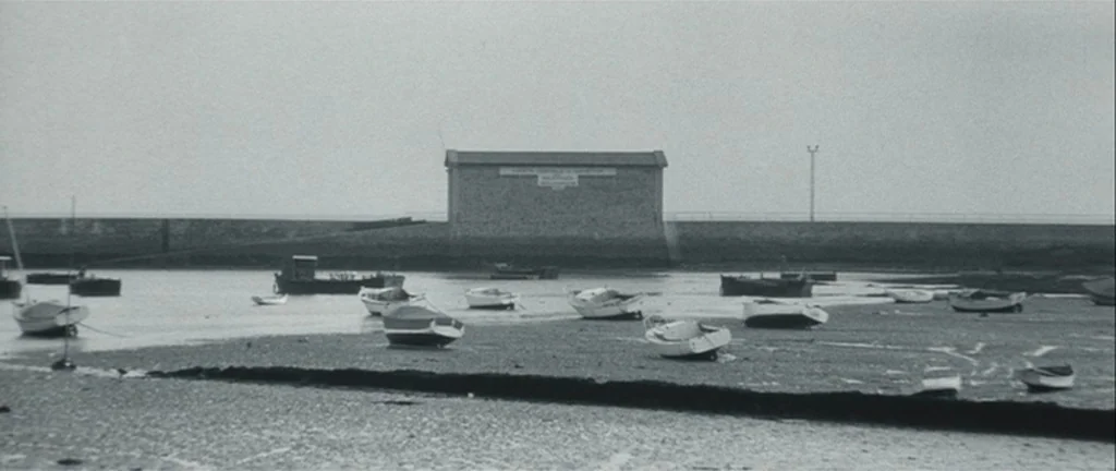 Les Créatures (1966) movie still: establishing — A tidal basin at low tide with numerous small boats resting on the wet sand. A stone pier and a…