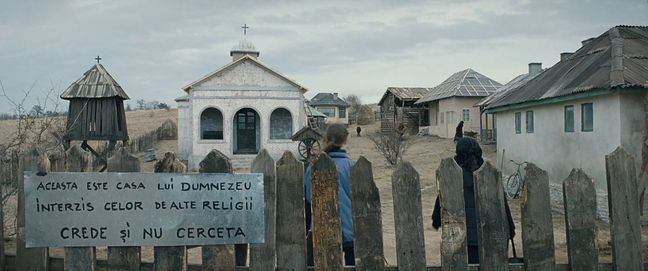 Beyond the Hills (2012) movie still: wide — A rural village features a small light-colored church with arched windows and wooden outbuildings. Two…