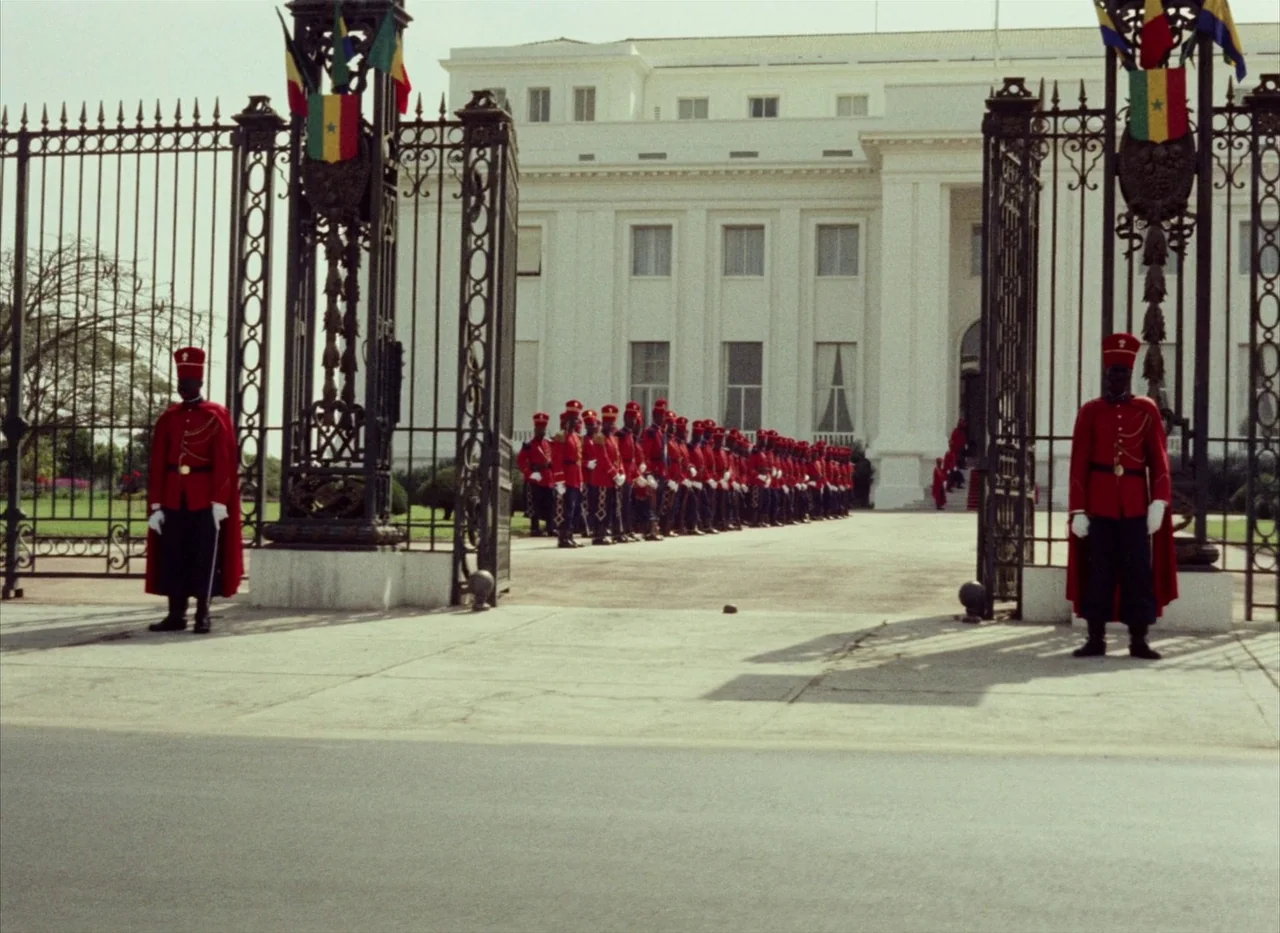 Touki Bouki (1973) movie still: wide — A grand white neoclassical building stands behind a black wrought iron entrance gate. A long line of soldiers…