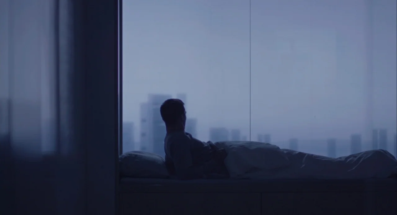 Equals (2015) movie still: wide — A modern bedroom overlooks a cityscape through a wall-sized glass window at dawn. A man reclines in bed and stares…