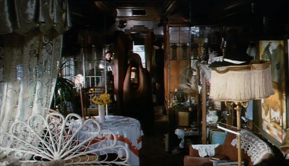 Harold and Maude (1971) movie still: wide — A cluttered interior space features ornate white wire furniture and lace curtains near a window. A tall…