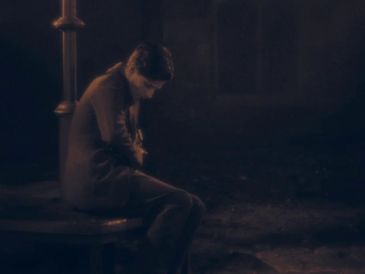 The Lodger (1927) movie still: medium — A dark city corner at night features a streetlamp illuminating a stone bench. A young man sits alone on the…