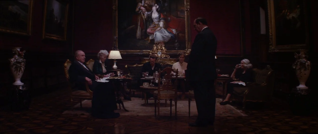 Neruda (2016) movie still: wide — A dimly lit, formal drawing room features ornate wood furniture, dark red walls, and oil paintings. Five people…