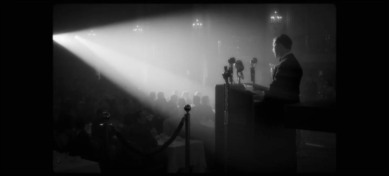 Mank (2020) movie still: medium — A dark banquet hall is illuminated by a single intense spotlight cutting through haze. A man in formal attire…