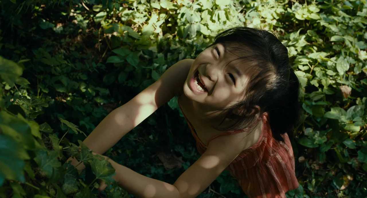 Last Summer (2023) movie still: medium — A dense patch of green ivy covers the ground under a sunlit tree canopy. A young East Asian girl in an…