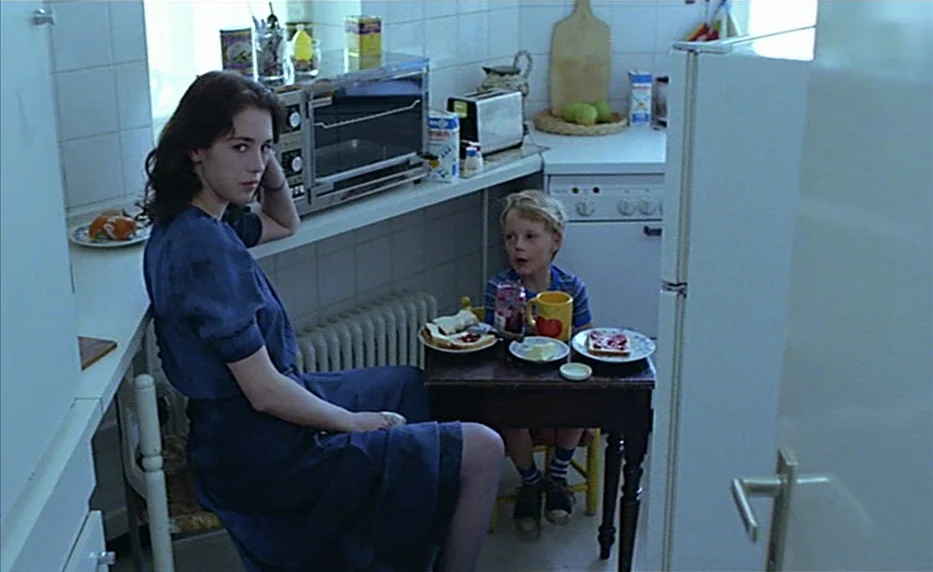 Possession (1981) movie still: medium — A cramped home space with white tiles and a small brown wooden dining table. A woman sits on a chair near a…