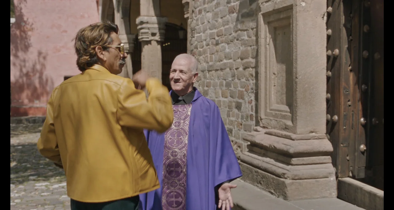 National Family (2023) movie still: medium — An outdoor courtyard with stone walls and an arched entryway sits under daylight. A man in a yellow…