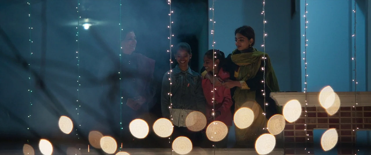The Wedding Guest (2018) movie still: fullbody — Three individuals stand on a balcony decorated with vertical hanging festive string lights. A woman…