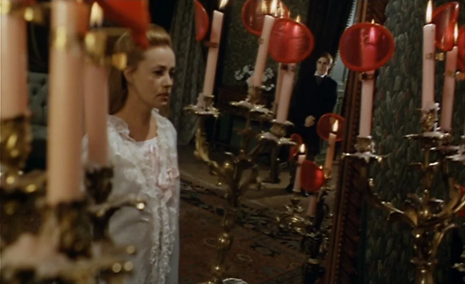 The Immortal Story (1968) movie still: medium — A dimly lit room features ornate candelabras with red accents and dark patterned wallpaper. A woman…