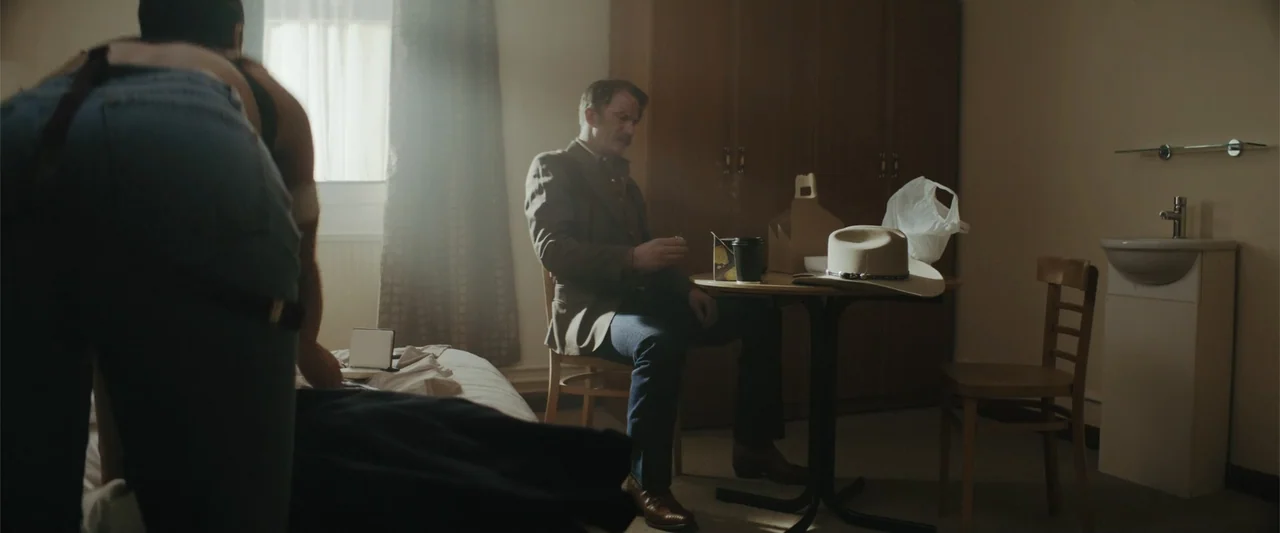 One Ranger (2023) movie still: medium — A sparse motel room interior features a small table with a white cowboy hat. A man sits at the table while a…
