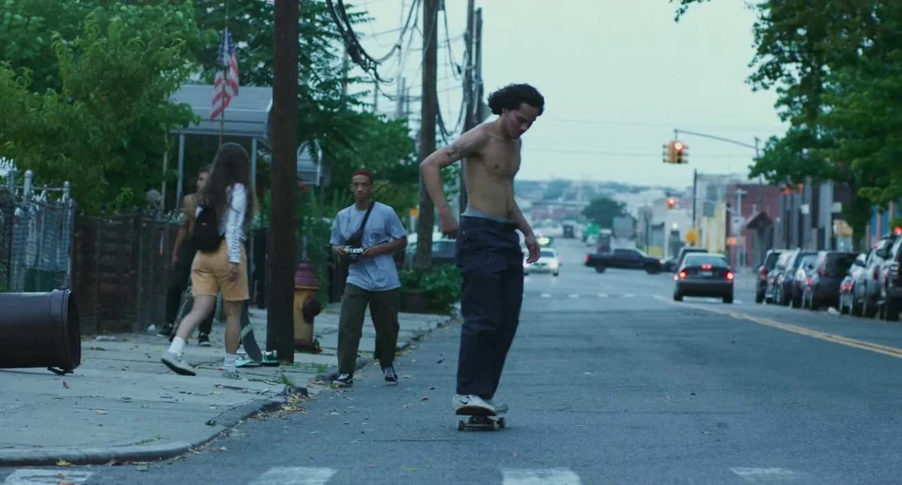 Skate Kitchen (2018) movie still: fullbody — A quiet urban street lined with parked cars and telephone poles on an overcast day. A young man…
