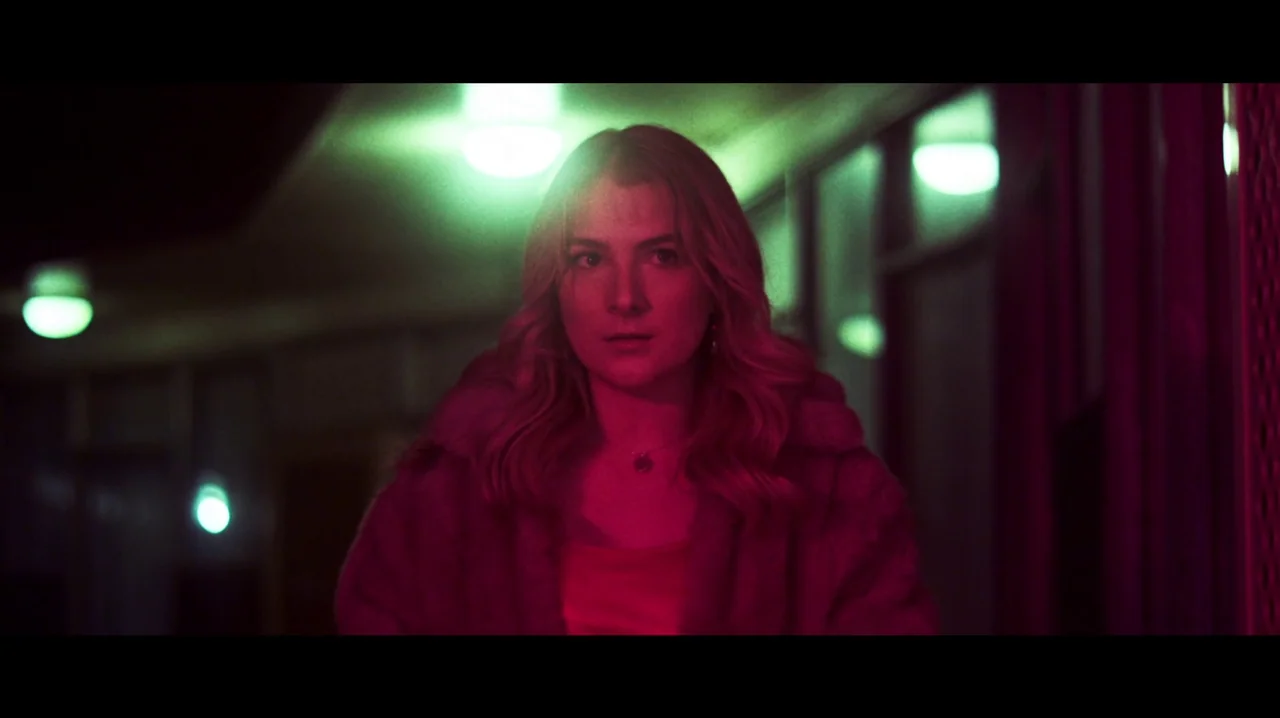 Carnage for Christmas (2024) movie still: medium — An exterior motel walkway at night featuring green and magenta artificial light. A young woman…