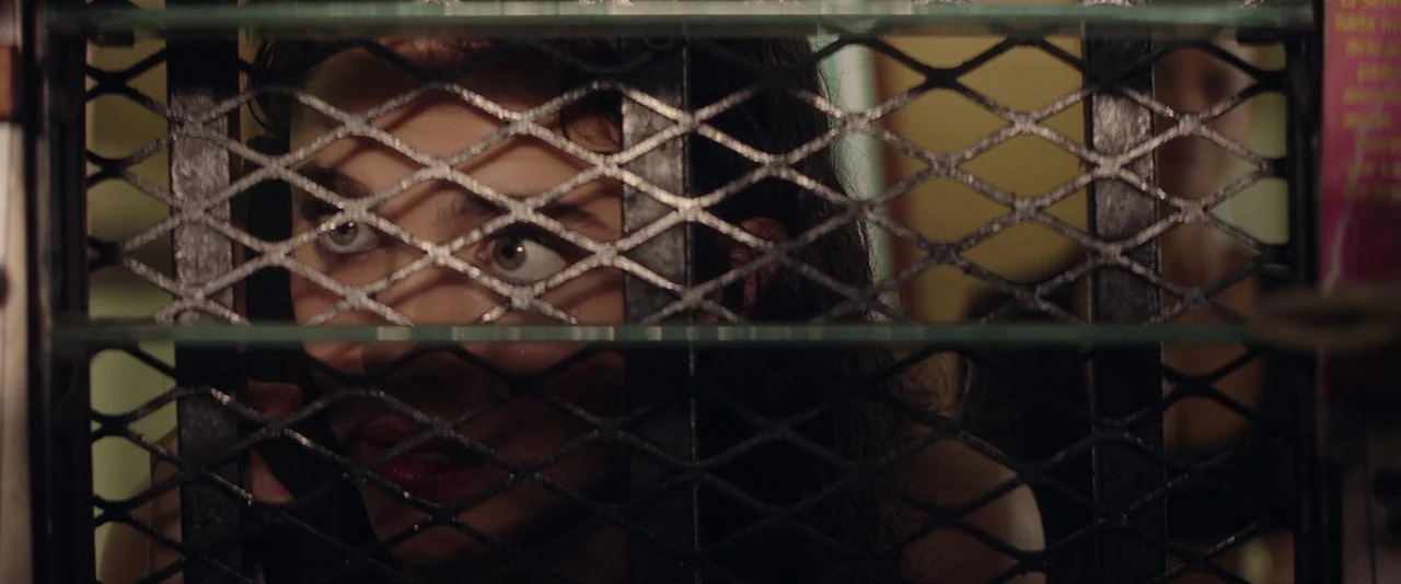 Stars At Noon (2022) movie still: closeup — A dark, partitioned space features a heavy black metal diamond-mesh security grill barrier. A woman…