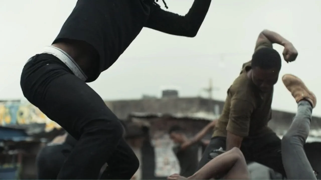 Gangs of Lagos (2023) movie still: medium — An outdoor area with weathered buildings and indistinct structures visible in the background. Two men…