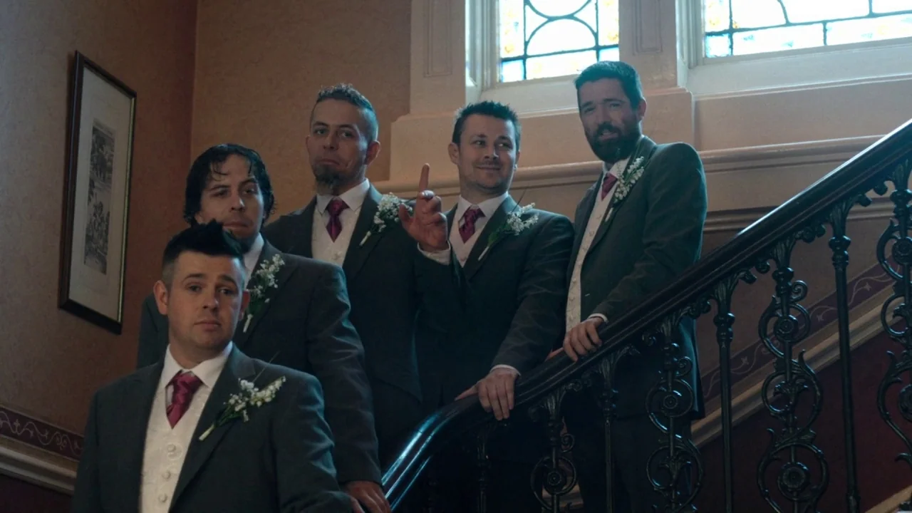 A Date for Mad Mary (2016) movie still: fullbody — Four men in formal wedding suits stand on an ornate indoor staircase. A tall stained glass window…