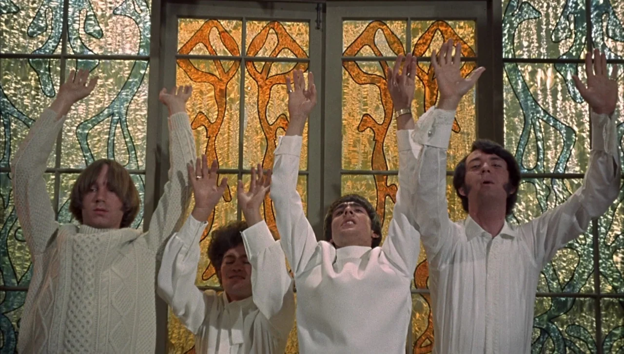 Head (1968) movie still: medium — Four men stand in front of large panels of amber-toned leaded stained glass windows. They hold their arms high…