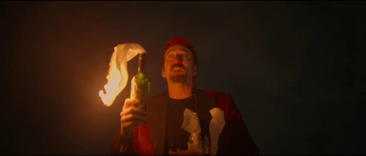 Sympathy For The Devil (2023) movie still: medium — A man stands against a dark, featureless background illuminated by a hand-held flame. He holds a…