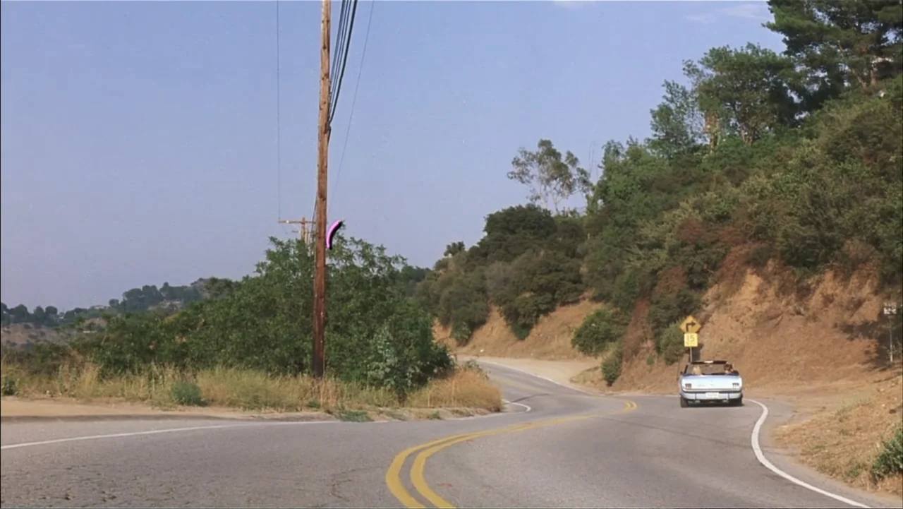 A Cinderella Story (2004) movie still: wide — A winding asphalt road curves through a hilly landscape with sparse trees and brush. A small silver…