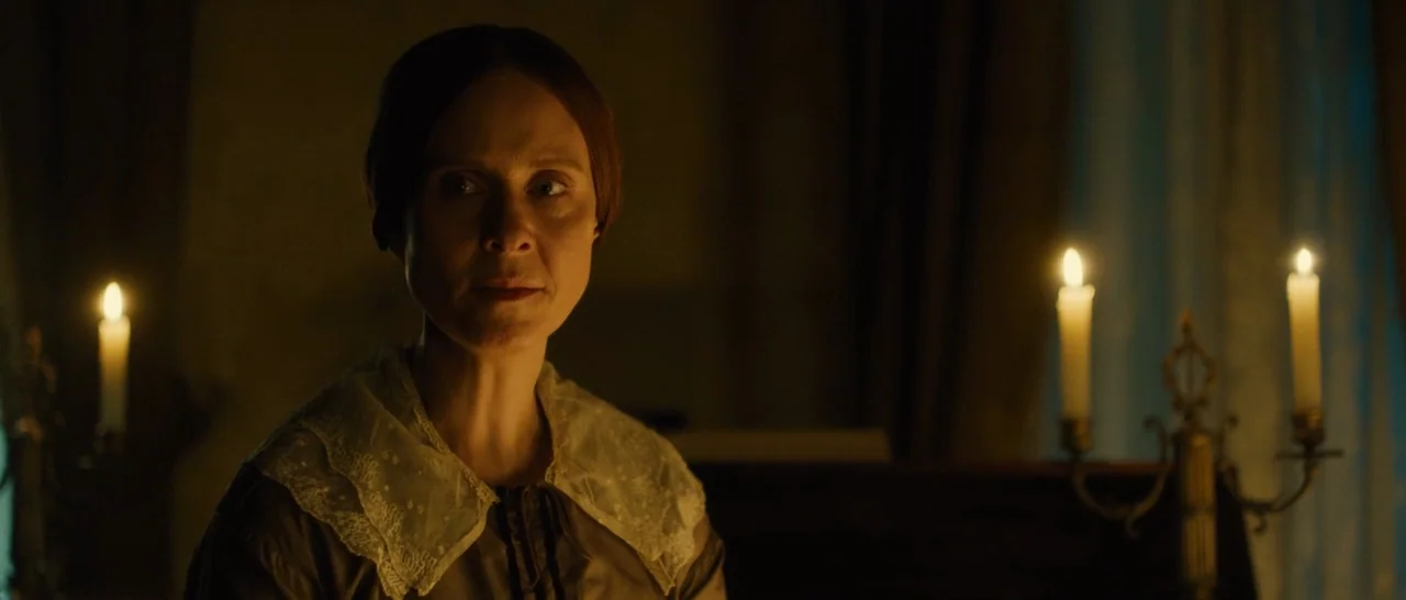 A Quiet Passion (2016) movie still: medium — A dimly lit 19th-century room featuring dark wood furniture and flickering candles. A woman with hair…