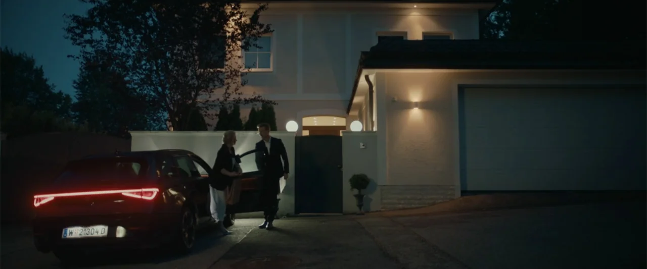 Peacock (2010) movie still: wide — A modern two-story house with a lit garage entrance faces a paved suburban driveway. A man and a woman stand by…