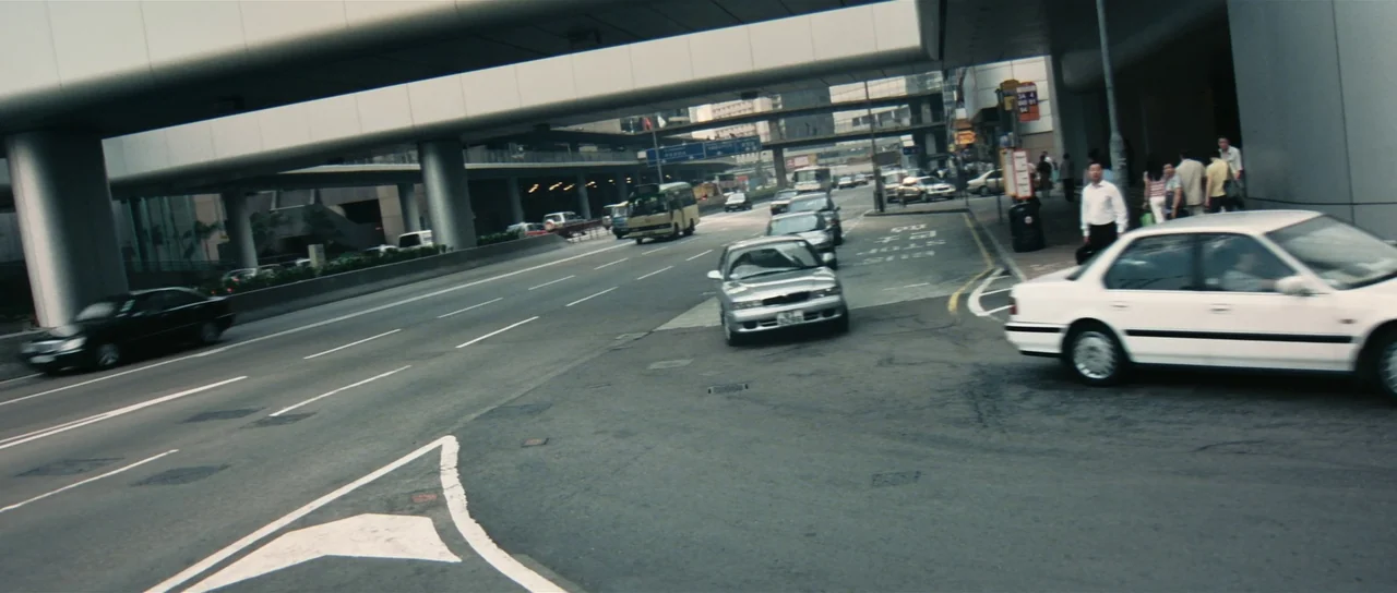 Infernal Affairs (2002) movie still: wide — A multi-lane asphalt road runs underneath a massive concrete overpass in an urban setting. Various cars…