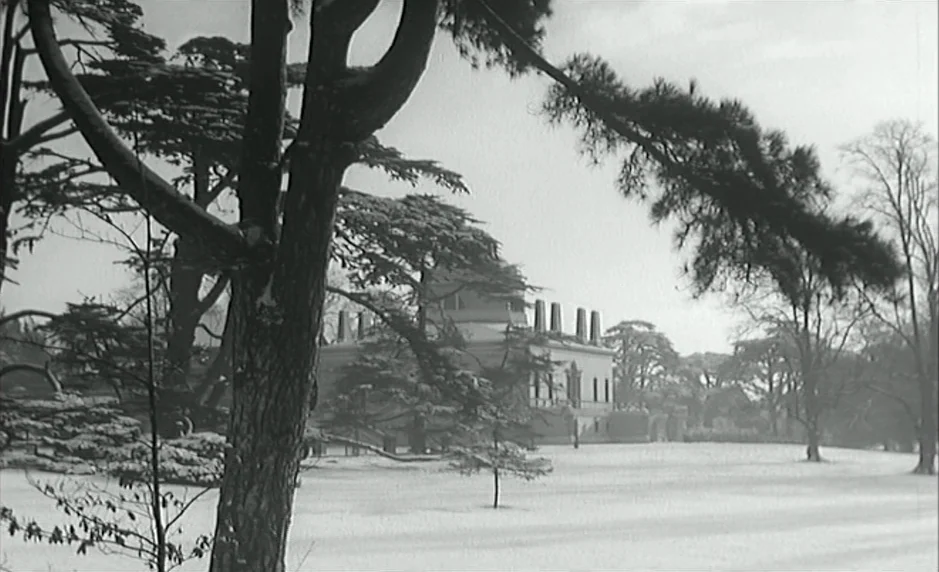 The Servant (1963) movie still: establishing — A snowy estate lawn is framed by large, dark tree branches in the foreground. A stately neoclassical…