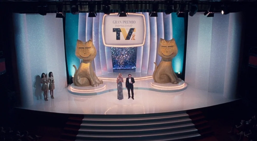 Somewhere (2010) movie still: wide — A wide television award show stage with two large gold stylized cat statues. Two presenters stand in the center…