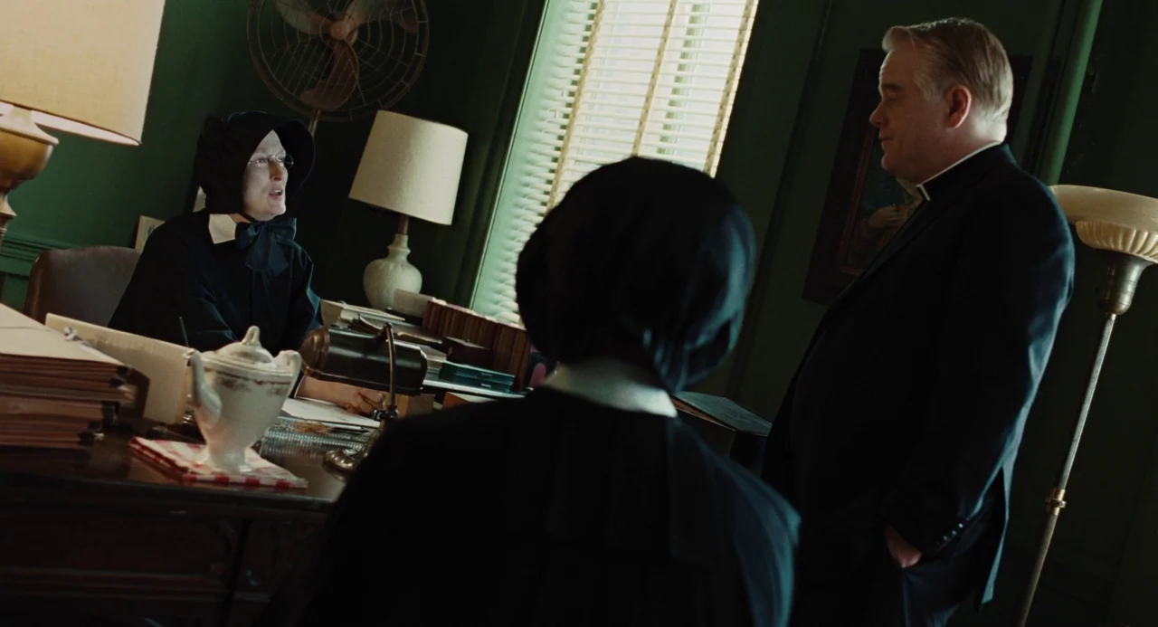 Doubt (2008) movie still: medium — An office with dark green walls, Venetian blinds, and a cluttered wooden desk. A nun sits behind the desk while a…
