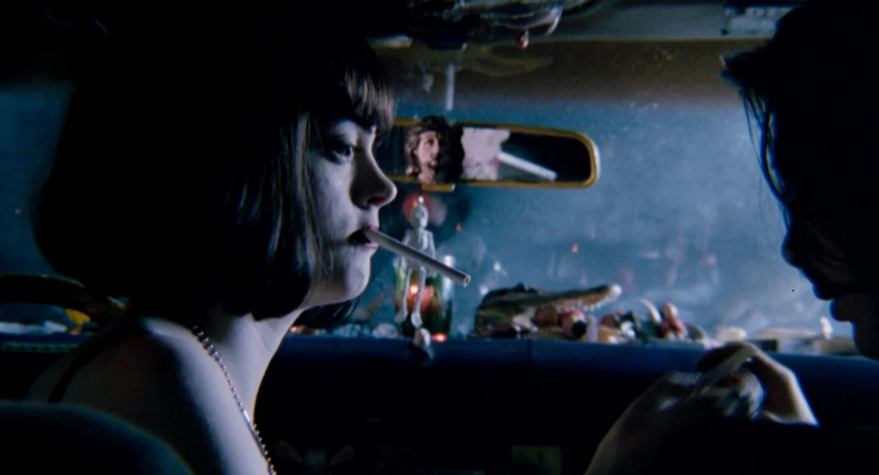 The Doom Generation (1995) movie still: medium — A dark car interior is filled with various dashboard ornaments including a skeleton figure. A woman…