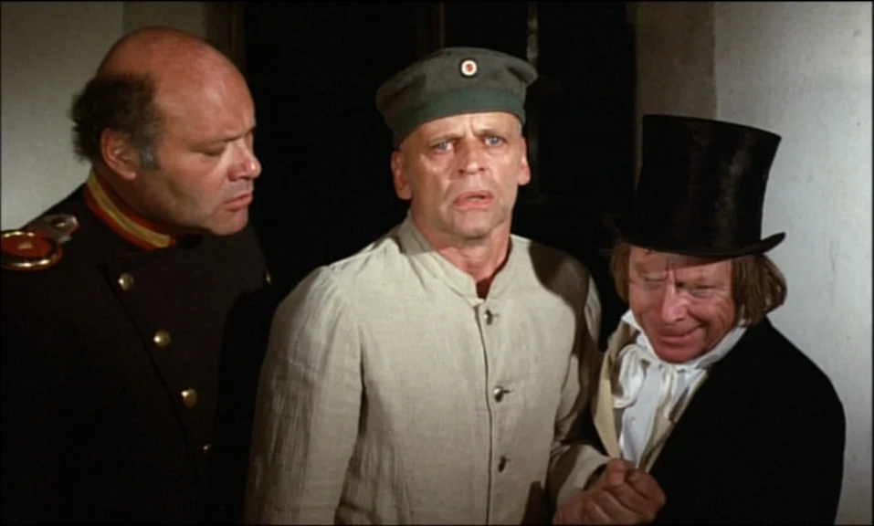 Woyzeck (1979) movie still: medium — A dark, dimly lit indoor space with plain walls serves as the backdrop. Three men in period clothing gather…