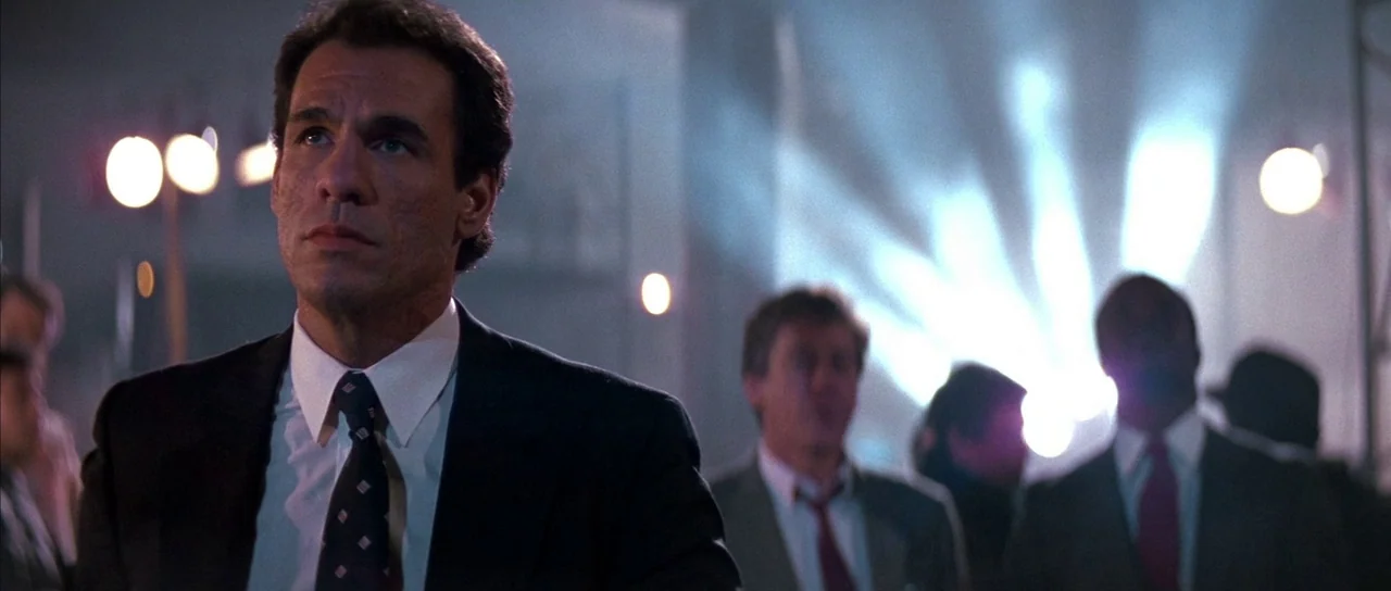 Die Hard (1988) movie still: medium — A dimly lit room features a man in a formal suit looking upward. Several other men in suits stand in the…