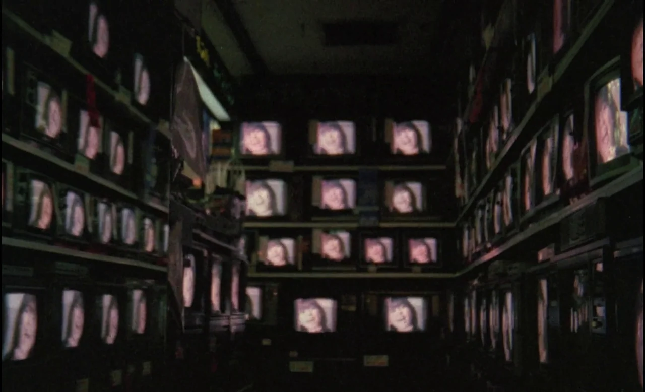 Sans Soleil (1983) movie still: establishing — Rows of vintage CRT television sets are stacked on shelves in a dark room. Each screen displays the…