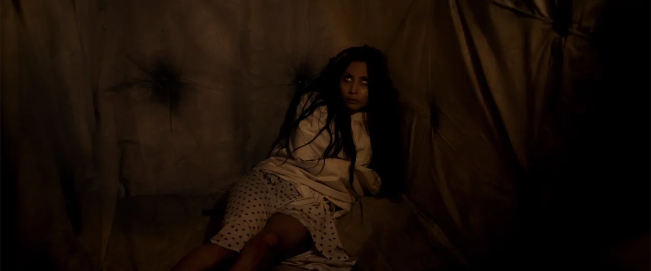 Mermaid Down (2019) movie still: medium — A dimly lit room with padded walls featuring tufted fabric surfaces. A young woman wears a straightjacket…