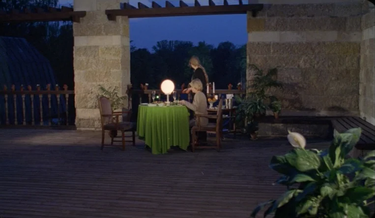 The Turn of the Century (2001) movie still: wide — A stone-walled terrace at twilight features a dining table covered in a green cloth. Two women…