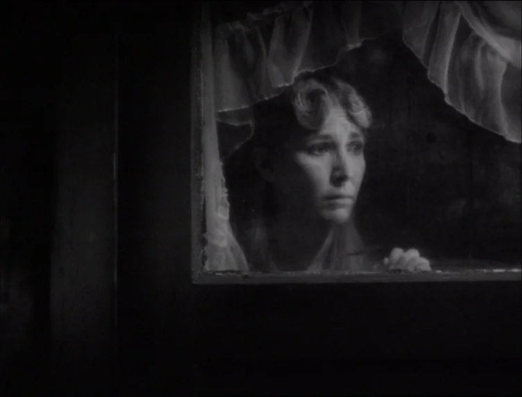 Eraserhead (1977) movie still: closeup — A woman stands behind a window with ruffled white curtains in a dark room. She peers through the glass with…