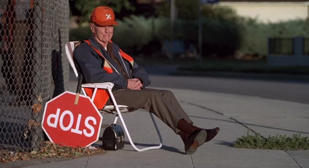 The Crossing Guard (1995) movie still: fullbody — An elderly man sits on a folding chair beside a chain-link fence on a sidewalk. He rests with a…