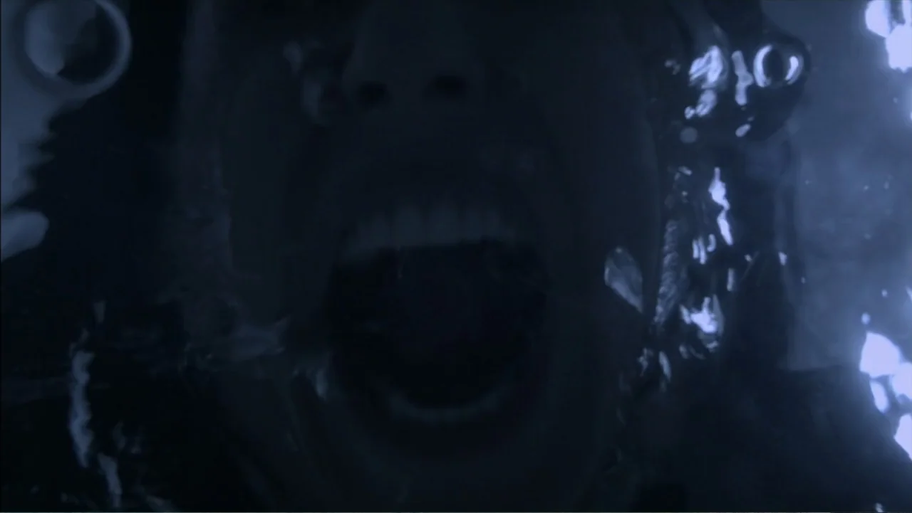 The Grotto (2022) movie still: closeup — An individual is submerged underwater in a dark, blue-lit environment with visible air bubbles. The person…